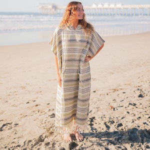 Puede incluir: Un poncho de playa con capucha con un patr&oacute;n de rayas verdes, grises y blancas. La prenda tiene un dobladillo con flecos y se usa en una playa de arena con el oc&eacute;ano al fondo.