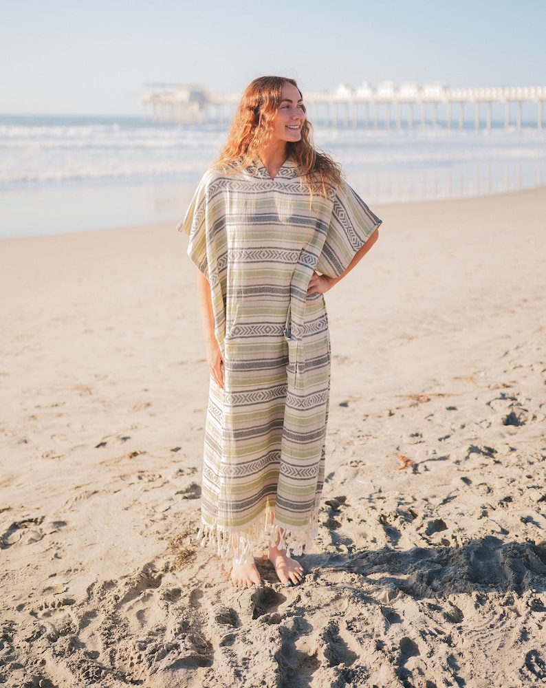 Puede incluir: Un poncho de playa con capucha con un patr&oacute;n de rayas verdes, grises y blancas. La prenda tiene un dobladillo con flecos y se usa en una playa de arena con el oc&eacute;ano al fondo.