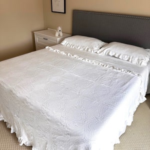 Op de afbeelding: Een witte sprei met een textuurpatroon en gefranjerde randen bedekt een bed. Twee witte kussens met gefranjerde randen liggen op het bed. Het hoofdbord is grijs met zilveren studs. Een wit nachtkastje en kunstwerken staan op de achtergrond.