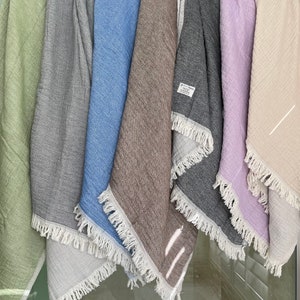 May include: A collection of six fringed throws in various colors, including green, gray, blue, brown, dark gray, lavender, and beige. The throws are made of a textured fabric and are hanging vertically.