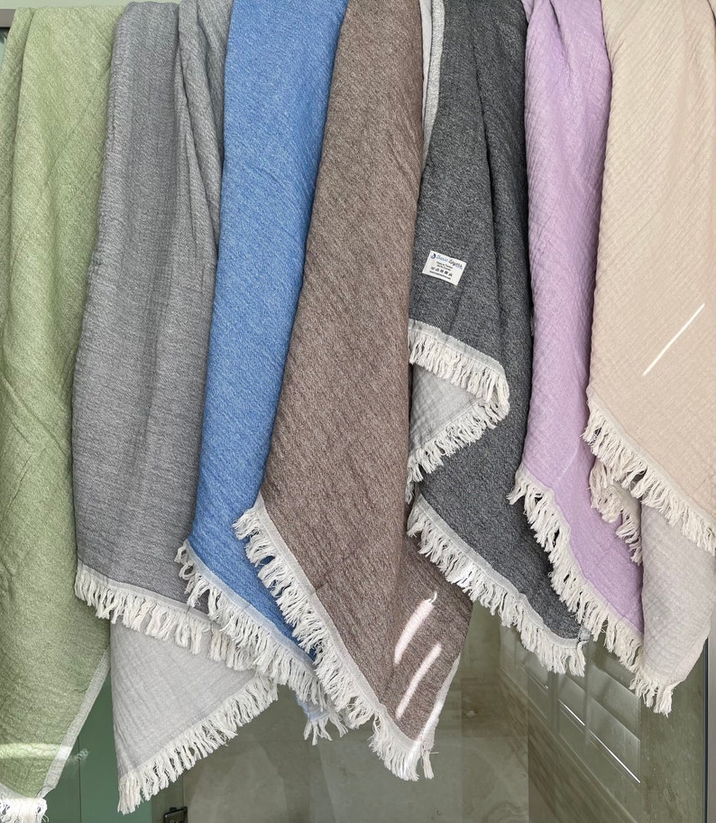 May include: A collection of six fringed throws in various colors, including green, gray, blue, brown, dark gray, lavender, and beige. The throws are made of a textured fabric and are hanging vertically.