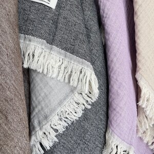 May include: Close-up of several fringed towels in various colors. A dark gray towel with white fringe is in the center, with a label that reads "Basic Layers." Other towels are light purple and beige. The towels are made of 100% cotton.