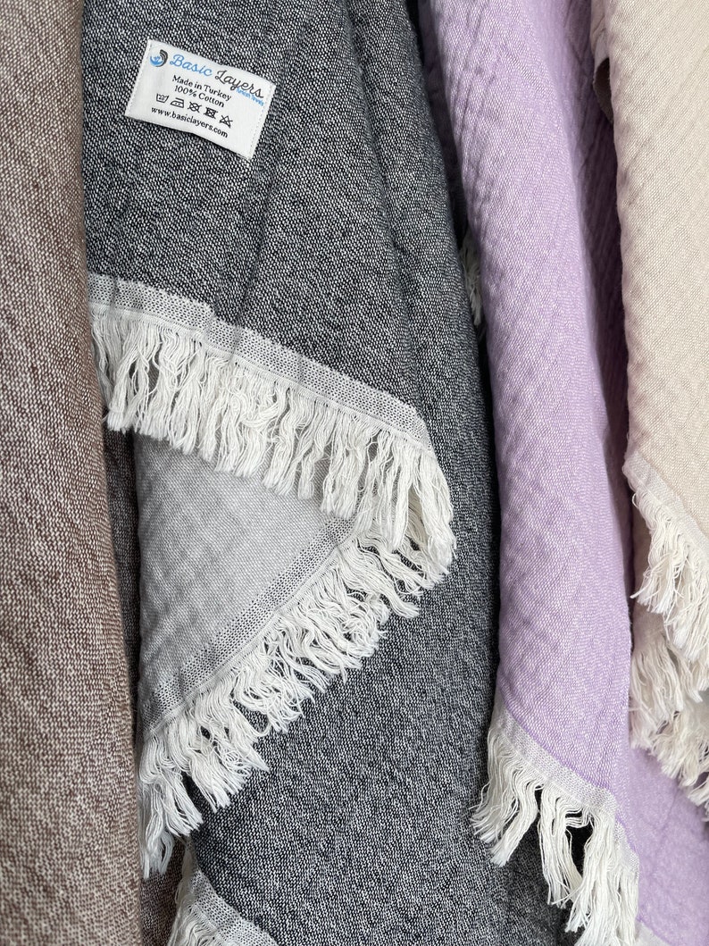 May include: Close-up of several fringed towels in various colors. A dark gray towel with white fringe is in the center, with a label that reads "Basic Layers." Other towels are light purple and beige. The towels are made of 100% cotton.