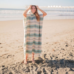 Puede incluir: Un poncho con capucha a rayas en tonos turquesa, beige y marr&oacute;n. El poncho tiene un borde con flecos y lo lleva una persona en una playa de arena, con el mar al fondo.