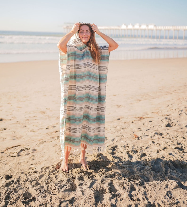 Puede incluir: Un poncho con capucha a rayas en tonos turquesa, beige y marr&oacute;n. El poncho tiene un borde con flecos y lo lleva una persona en una playa de arena, con el mar al fondo.