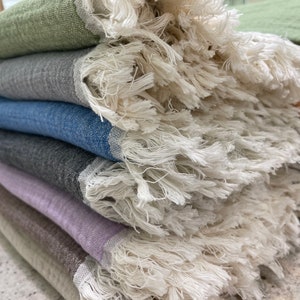May include: A stack of colorful fringed throws in shades of green, gray, blue, and purple. The throws have a textured weave and a thick, off-white fringe along the edges. The throws are neatly folded, showcasing the variety of colors and textures.