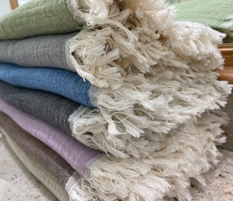 May include: A stack of colorful fringed throws in shades of green, gray, blue, and purple. The throws have a textured weave and a thick, off-white fringe along the edges. The throws are neatly folded, showcasing the variety of colors and textures.