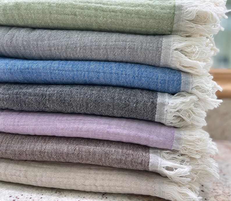 May include: A stack of colorful, textured towels in shades of green, gray, blue, black, lavender, brown, and beige. Each towel has a fringed edge in white.