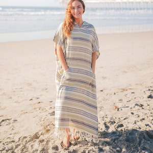 Puede incluir: Una larga cubierta de playa con capucha con un estampado de rayas en tonos verdes, grises y blancos. La cubierta tiene un dobladillo con flecos y dos bolsillos delanteros. La persona est&aacute; de pie en una playa de arena con el oc&eacute;ano y un muelle al fondo.