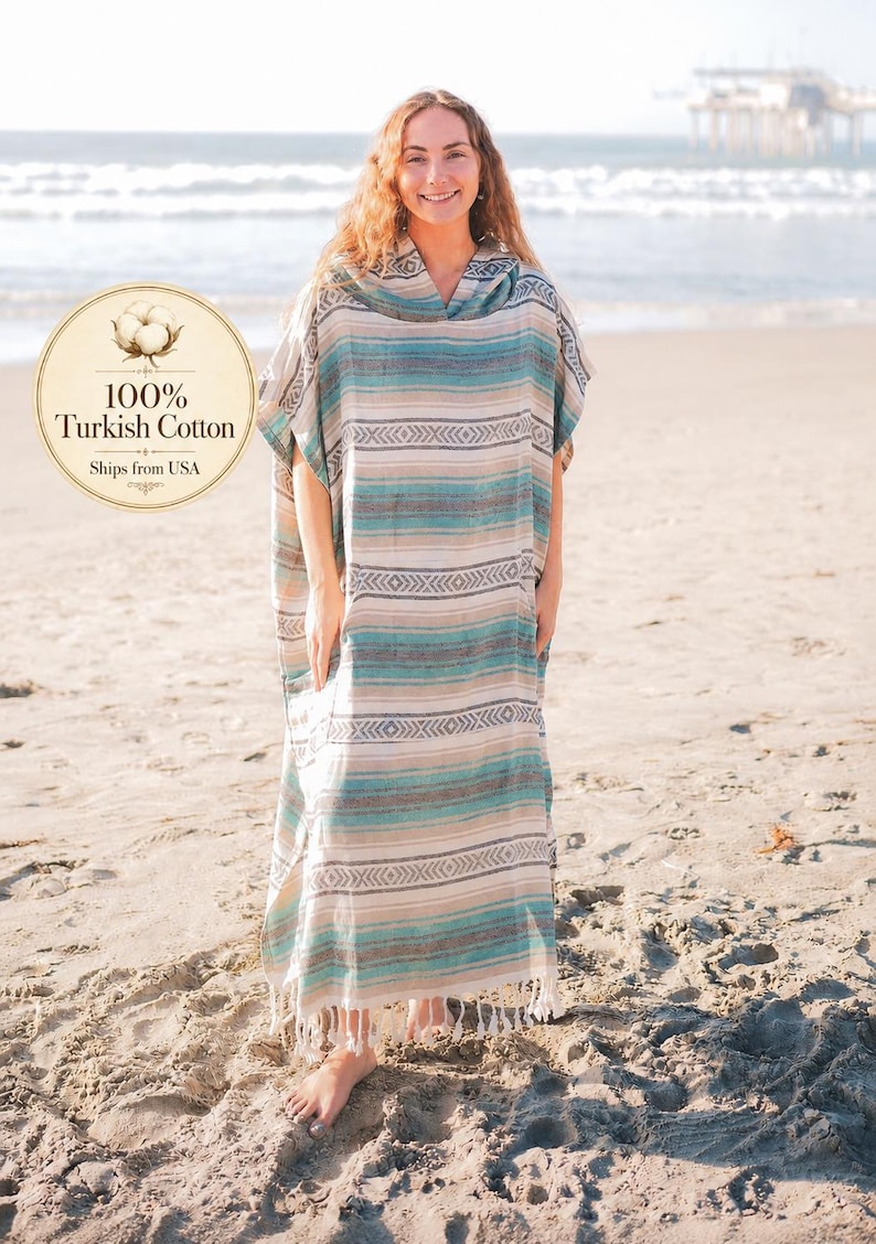 Puede incluir: Una mujer con un poncho de playa largo y a rayas con capucha, de pie en una playa de arena. El poncho presenta rayas horizontales en tonos azules, beige y blanco. El texto "100% Turkish Cotton" es visible.