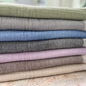 May include: A stack of folded linen throws in various colors, including sage green, gray, blue, charcoal, lavender, brown, and cream. Each throw has a fringed edge. The throws are made of a textured fabric.