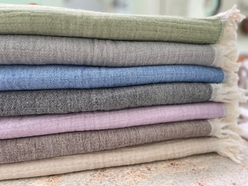 May include: A stack of folded linen throws in various colors, including sage green, gray, blue, charcoal, lavender, brown, and cream. Each throw has a fringed edge. The throws are made of a textured fabric.