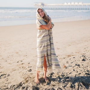 Puede incluir: Poncho de playa con capucha con un estampado de rayas en tonos verdes, blancos y grises. El poncho tiene una abertura frontal y flecos a lo largo del dobladillo inferior. La persona est&aacute; de pie en una playa de arena cerca del oc&eacute;ano.