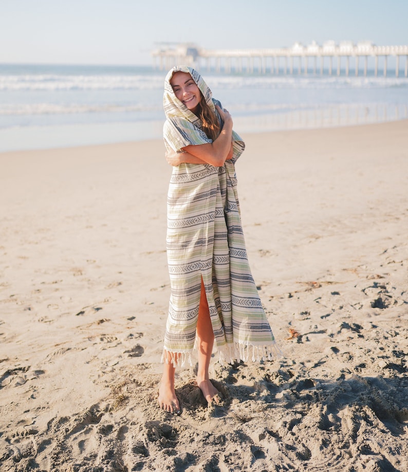 Puede incluir: Poncho de playa con capucha con un estampado de rayas en tonos verdes, blancos y grises. El poncho tiene una abertura frontal y flecos a lo largo del dobladillo inferior. La persona est&aacute; de pie en una playa de arena cerca del oc&eacute;ano.