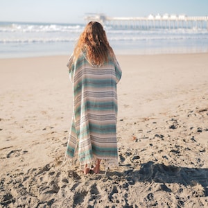 Puede incluir: Un poncho de playa a rayas en tonos turquesa, beige y blanco, con capucha y dobladillo con flecos. El poncho se lleva en una playa de arena, con el mar y un muelle al fondo.