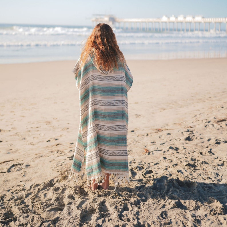Puede incluir: Un poncho de playa a rayas en tonos turquesa, beige y blanco, con capucha y dobladillo con flecos. El poncho se lleva en una playa de arena, con el mar y un muelle al fondo.