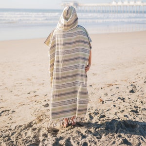 Puede incluir: Un poncho con capucha con un patr&oacute;n de rayas en tonos verdes, grises y crema. El poncho tiene un dobladillo con flecos y se usa en una playa de arena con el oc&eacute;ano y un muelle al fondo.