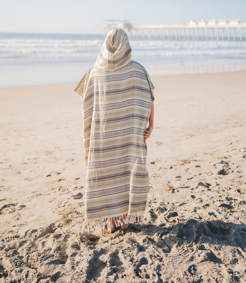 Puede incluir: Un poncho con capucha con un patr&oacute;n de rayas en tonos verdes, grises y crema. El poncho tiene un dobladillo con flecos y se usa en una playa de arena con el oc&eacute;ano y un muelle al fondo.