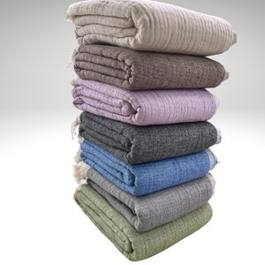 May include: A stack of seven folded blankets in various colors, including beige, brown, lavender, gray, blue, and green. Each blanket has a textured appearance and is neatly folded, with visible fringe on some. The blankets are stacked on top of each other.