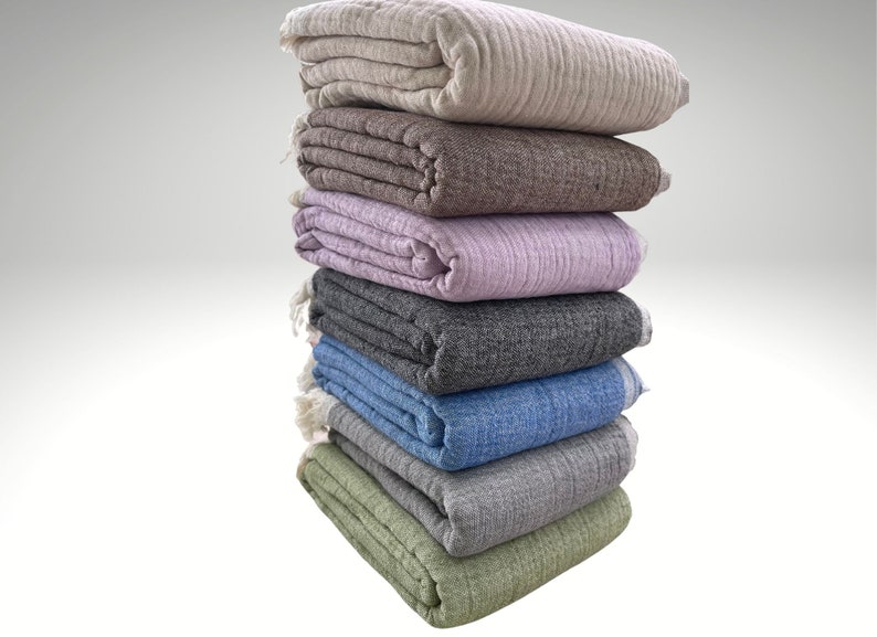 May include: A stack of seven folded blankets in various colors, including beige, brown, lavender, gray, blue, and green. Each blanket has a textured appearance and is neatly folded, with visible fringe on some. The blankets are stacked on top of each other.