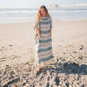 Puede incluir: Un poncho largo con capucha con un estampado de rayas en tonos azules, beige y blanco. El poncho tiene bolsillos y un dobladillo con flecos. La persona est&aacute; de pie en una playa de arena cerca del oc&eacute;ano.