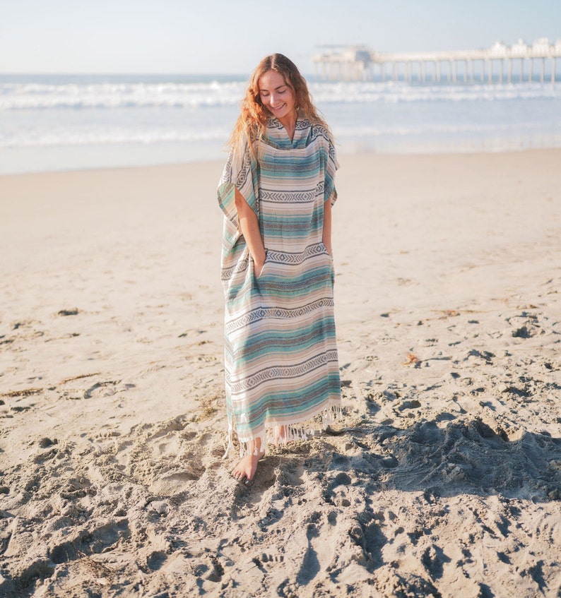 Puede incluir: Un poncho largo con capucha con un estampado de rayas en tonos azules, beige y blanco. El poncho tiene bolsillos y un dobladillo con flecos. La persona est&aacute; de pie en una playa de arena cerca del oc&eacute;ano.