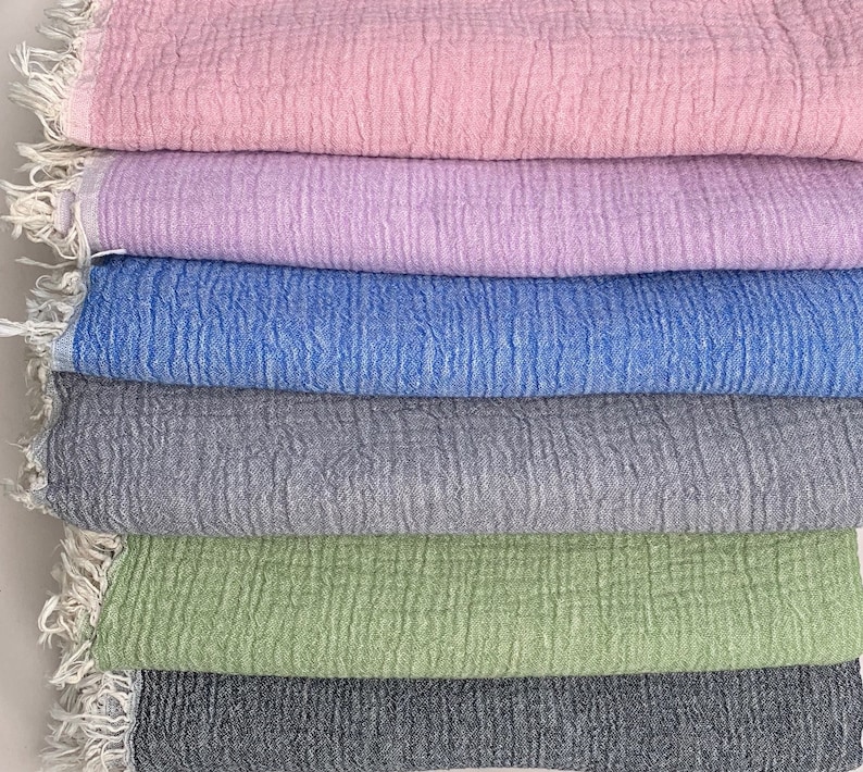 May include: A stack of seven cotton towels in various pastel colors. The towels are pink, lavender, blue, gray, green, and charcoal. Each towel has a fringed edge.