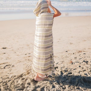 Puede incluir: Una t&uacute;nica de playa con capucha y estampado de rayas en tonos verdes, blancos y grises. La t&uacute;nica tiene un dise&ntilde;o largo y fluido con una abertura lateral y flecos en el bajo. La persona est&aacute; en una playa de arena con el mar al fondo.