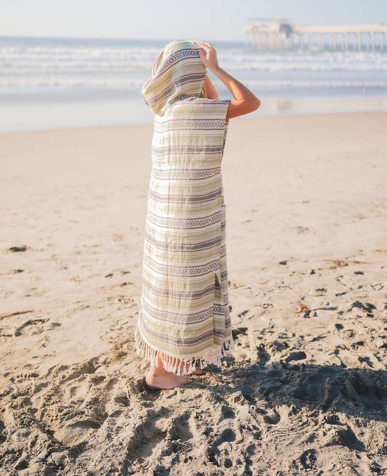 Puede incluir: Una t&uacute;nica de playa con capucha y estampado de rayas en tonos verdes, blancos y grises. La t&uacute;nica tiene un dise&ntilde;o largo y fluido con una abertura lateral y flecos en el bajo. La persona est&aacute; en una playa de arena con el mar al fondo.
