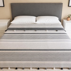 May include: A gray and white striped blanket with tassels, displayed on a bed. The blanket features varying shades of gray and white horizontal stripes. Decorative tassels are along the bottom edge. The bed has white pillows and a gray headboard.