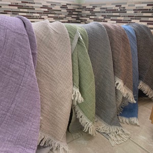 May include: A collection of soft, textured throws in various colors, including lavender, beige, green, gray, brown, and blue. Each throw has a fringed edge. The throws are displayed in a row, showcasing their different hues and textures.