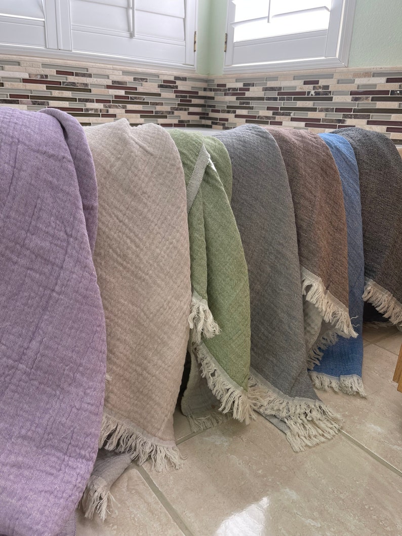 May include: A collection of soft, textured throws in various colors, including lavender, beige, green, gray, brown, and blue. Each throw has a fringed edge. The throws are displayed in a row, showcasing their different hues and textures.
