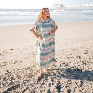 Puede incluir: Un poncho largo con capucha con un estampado de rayas en tonos azules, verdes y beige. El poncho tiene un dobladillo con flecos y se usa en una playa de arena con el oc&eacute;ano al fondo.
