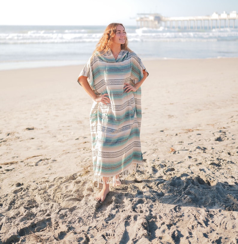 Puede incluir: Un poncho largo con capucha con un estampado de rayas en tonos azules, verdes y beige. El poncho tiene un dobladillo con flecos y se usa en una playa de arena con el oc&eacute;ano al fondo.