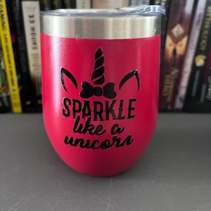 Puede incluir: Un vaso térmico rojo con borde y tapa plateados. El vaso presenta un diseño de unicornio negro y las palabras "SPARKLE like a unicorn" en escritura negra. El vaso es cilíndrico y se apoya sobre una superficie gris.