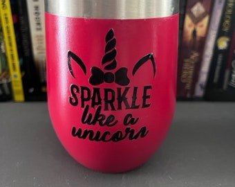 May include: A red insulated tumbler with a silver rim and lid. The tumbler features a black unicorn design and the words "SPARKLE like a unicorn" in black script. The tumbler is cylindrical and sits on a gray surface.