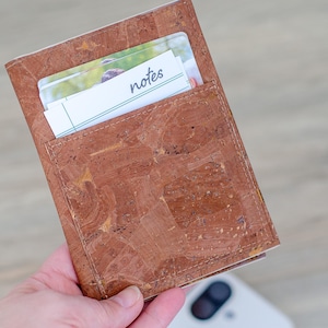May include: A brown cork wallet with a pocket holding a few white cards. The word "notes" is visible on one of the cards. The wallet is held in a hand.