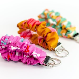 May include: Three colourful fabric keychains with metal key rings. The keychains are made of fabric with a ruffled design and feature different floral patterns. The keychains are pink, orange, and green.