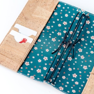 Könnte beinhalten: Offenes Notizbuch aus Kork mit Kartenfächern. Der Innenraum hat einen teal-farbenen Blumenstoff mit weißen und rosa Blüten. Schwarze Gummibänder sind sichtbar. Zwei Karten befinden sich in den Kartenfächern, eine mit einer roten Handgrafik.