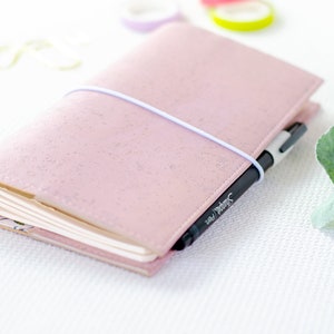 Könnte beinhalten: Ein rosa Notizbuch mit strukturierter Hülle, durch ein weißes Gummiband verschlossen. Ein schwarzer Sharpie-Stift steckt im Buchrücken. Die Seiten des Journals sind sichtbar, und das Innenfutter hat ein Blumenmuster.
