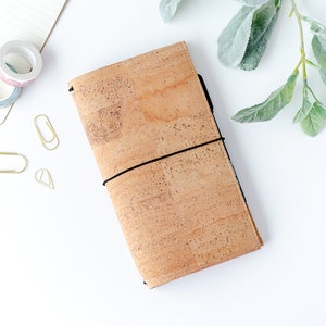 May include: A cork journal with a black elastic closure, on a white surface. Decorative washi tape rolls and gold paper clips are nearby. A sprig of green foliage is in the upper right corner. The journal is approximately 20 cm long.