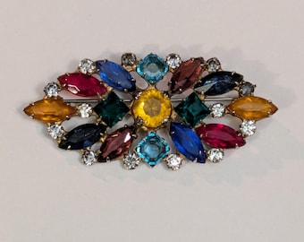 May include: A gold-toned brooch with a cluster of colorful rhinestones in various shapes and sizes. The rhinestones are arranged in a symmetrical pattern, with a central yellow rhinestone surrounded by blue, green, red, and orange rhinestones.