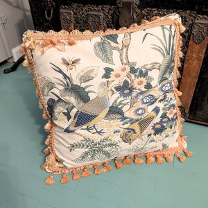 May include: Square decorative pillow with a cream background featuring a bird and floral design in blue, green, and yellow. The pillow has a peach-colored trim with tassels around the edges. The pillow is placed on a light blue surface.