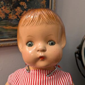 May include: A vintage porcelain doll with a red and white striped dress and a silver collar. The doll has blonde hair, blue eyes, and a painted smile.