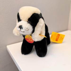 May include: A plush black and white dog toy with a cream-colored head and ears. The toy has a yellow tag with red text that reads "Cockle". A second yellow tag is also attached. The toy is sitting on a white surface.