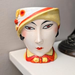 May include: A vintage ceramic head vase with a stylized face, featuring red lips and eyes, and blush on the cheeks. The vase has a yellow and white hat with red accents and a red bow. The face is painted with black eyeliner and eyebrows.