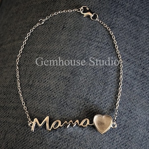 May include: A silver bracelet with the word "Mama" in cursive script and a heart-shaped charm. The bracelet is made of delicate chain links and has a clasp closure. The image is set against a dark denim background.