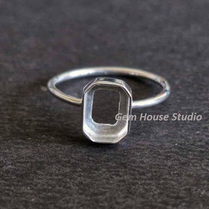 Może przedstawiać: Srebrny pierścionek z oprawą o szlifie szmaragdowym. Pierścionek jest prezentowany na ciemnej powierzchni. Na pierścionku widoczny jest napis "Gem House Studio".