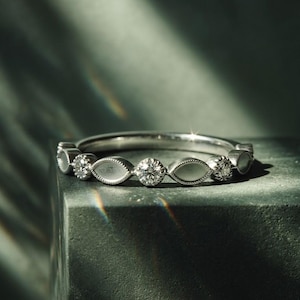 May include: A silver ring with a series of round and marquise-shaped settings, each holding a sparkling stone. The ring is set against a dark, textured background, with light creating a subtle highlight on the metal and stones. A piece of jewelry.