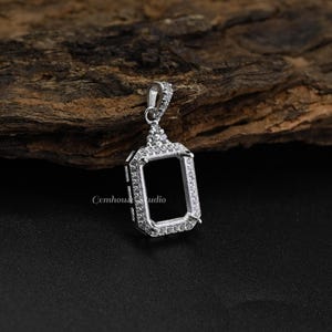 May include: A silver pendant setting with a rectangular centre, encircled by small, clear gemstones. The setting has a decorative bail and is designed to hold a gemstone. The text "Gemhouse Studio" is visible on the setting.